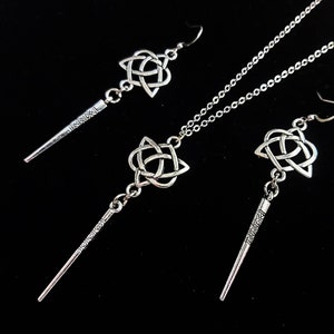 May include: A silver jewellery set comprising a necklace and two earrings. Each item features a Celtic knot design and a wand-shaped pendant with etched detailing. The set is presented against a black backdrop.