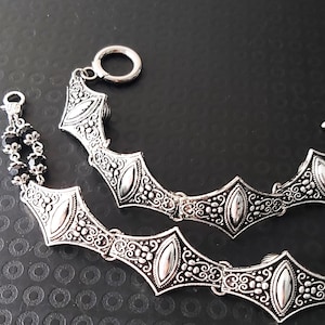 May include: Two silver bracelets with detailed, geometric patterns. Each bracelet has a series of oval-shaped elements linked by intricate designs. One bracelet has a toggle clasp, the other a lobster clasp and black bead accents. The bracelets are shown on a dark surface.