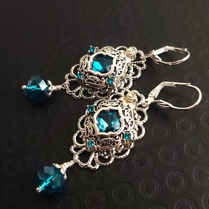 May include: A pair of ornate silver-toned earrings with teal-colored gemstones. Each earring features a filigree design with a large square gem in the center and smaller gems around it. A faceted teal bead dangles from the bottom. The earrings have a lever-back closure.