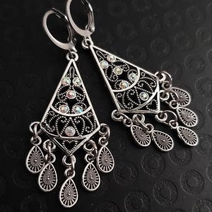 May include: A pair of silver-toned drop earrings featuring a triangular filigree frame with embedded iridescent crystals. The earrings hang from lever-back closures and include multiple small, textured teardrop-shaped charms at the base.