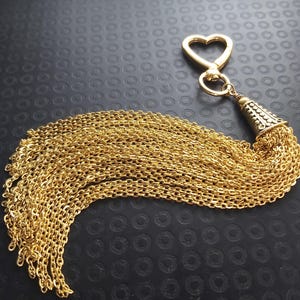 May include: Gold-colored keychain with a heart-shaped clasp and a decorative tassel made of multiple chains. The tassel is attached to a cone-shaped element with a detailed pattern. The background is black.