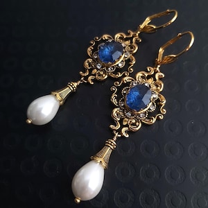 Peut inclure: Une paire de boucles d'oreilles dorées ornées. Chaque boucle d'oreille présente une grande pierre précieuse ovale bleu foncé entourée de petites pierres claires. En dessous, une perle blanche en forme de goutte. Design vintage et élégant.