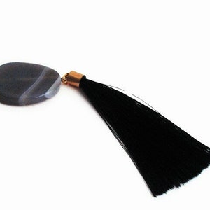 Op de afbeelding: Een sleutelhanger met een grijze stenen hanger met witte aderen, een zwarte kwast en een goudkleurige sluiting. De steen is ovaal van vorm. De kwast is lang en gemaakt van zwarte draden.