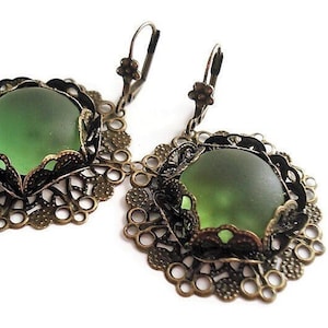 May include: A pair of antique-style earrings featuring large, round, frosted green glass stones set in ornate, filigree metalwork. The earrings have a lever-back closure and a bronze-colored finish, creating a vintage aesthetic. The design is symmetrical and detailed.