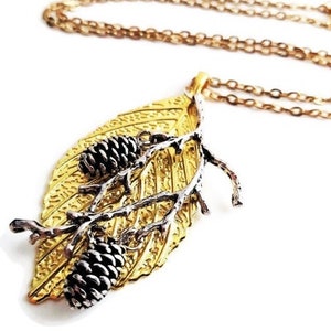 Puede incluir: Un collar con un colgante de hoja dorada, una rama plateada y dos piñas. El collar está en una cadena dorada.