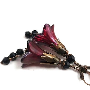 May include: A pair of dangle earrings featuring two red flower charms and black beads. The earrings have a bronze-coloured metal base.