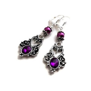Puede incluir: Un par de pendientes colgantes plateados con un diseño vintage. Cada pendiente presenta una gema grande, ovalada, de color púrpura oscuro, rodeada de filigranas ornamentadas y acentuada con cuentas púrpuras más pequeñas.