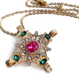 Puede incluir: Un collar de oro con un colgante que presenta una gran piedra preciosa rosa rodeada de piedras preciosas transparentes más pequeñas y cuatro piedras preciosas verdes. El colgante tiene la forma de una estrella.