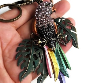May include: A jeweled parrot keychain with a green, yellow, pink, and purple tail. The parrot is surrounded by green leaves.