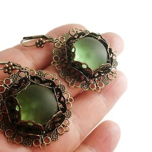 May include: A pair of vintage-style earrings featuring olive green glass stones set in ornate, filigree bronze-toned metal frames. The earrings have a lever-back closure and a delicate, floral design around the edges. The earrings are a unique accessory.
