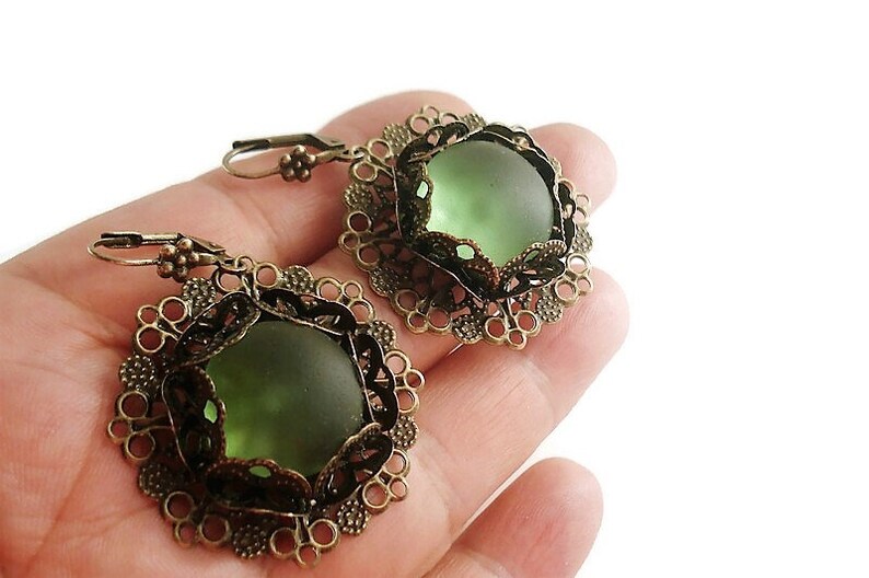 May include: A pair of vintage-style earrings featuring olive green glass stones set in ornate, filigree bronze-toned metal frames. The earrings have a lever-back closure and a delicate, floral design around the edges. The earrings are a unique accessory.