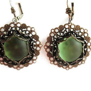 May include: A pair of earrings featuring olive green glass stones set in ornate, bronze-colored filigree frames. The earrings have a lever-back closure and a delicate, vintage aesthetic. The filigree design is circular with intricate detailing.