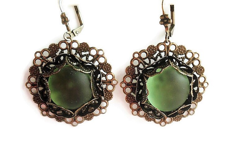 May include: A pair of earrings featuring olive green glass stones set in ornate, bronze-colored filigree frames. The earrings have a lever-back closure and a delicate, vintage aesthetic. The filigree design is circular with intricate detailing.