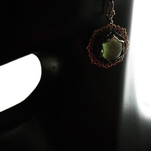 May include: A bronze-colored earring with a circular design and a central, light green stone. The earring is displayed on a black mannequin, showcasing its intricate details and craftsmanship. The design features a floral-like pattern.