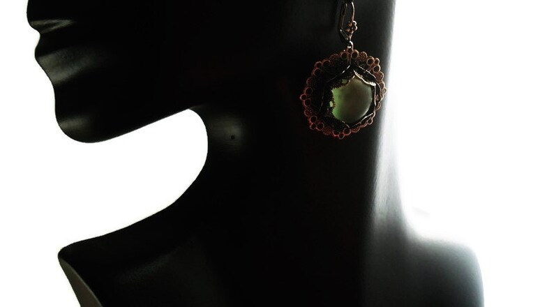 May include: A bronze-colored earring with a circular design and a central, light green stone. The earring is displayed on a black mannequin, showcasing its intricate details and craftsmanship. The design features a floral-like pattern.