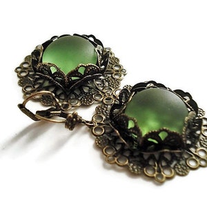 May include: A pair of vintage-style earrings featuring a round, frosted green glass stone set in an ornate, filigree bronze-toned metal frame. The earrings have a lever-back closure and are displayed on a white background.