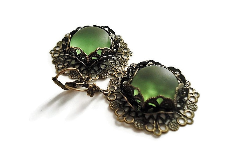 May include: A pair of vintage-style earrings featuring a round, frosted green glass stone set in an ornate, filigree bronze-toned metal frame. The earrings have a lever-back closure and are displayed on a white background.