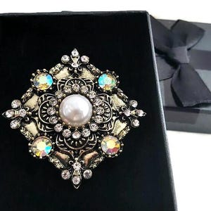 May include: A gold-toned brooch with a square design featuring a large faux pearl surrounded by smaller clear rhinestones and iridescent stones.