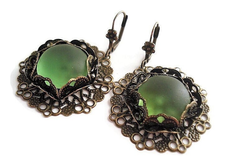 May include: A pair of antique-style earrings featuring large, round, frosted green glass stones set in ornate, filigree metalwork. The earrings have a lever-back closure and a bronze-colored finish, creating a vintage aesthetic. The design is symmetrical and detailed.