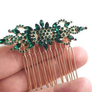 May include: A gold hair comb with a large emerald green rhinestone design. The comb has a decorative floral pattern with a central cluster of rhinestones.
