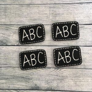 May include: Four black felt patches with white stitching around the edges. Each patch has the letters "ABC" embroidered in white.