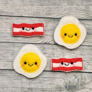 May include: Two sets of felt appliques in the shape of a fried egg and a strip of bacon. Each applique has a smiling face embroidered on it. The appliques are white, yellow, and red.