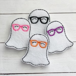 May include: Four white felt ghost-shaped ornaments with black stitched outlines. Each ghost wears a different color of glasses: black, pink, orange, and purple. The ghosts are arranged on a white wooden surface.