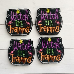 May include: Four black felt patches shaped like cauldrons, each embroidered with the purple, green, and orange text Witch in Training, a yellow star, and two small pink spiders. Each patch features a white stitched border around the edge.
