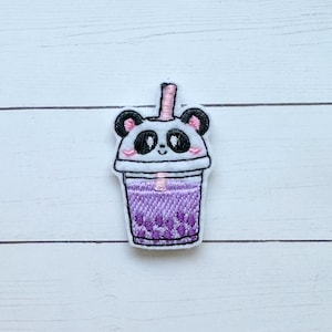 May include: Embroidered patch featuring a panda bear design atop a purple bubble tea cup. The panda has black ears, eyes, and a pink blush. The cup has a pink straw and a white rim. The patch is on a white wooden surface.
