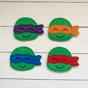 May include: Four felt appliques of Teenage Mutant Ninja Turtles. Each turtle has a green head with a different colored eye mask. The masks are purple, orange, blue, and red.