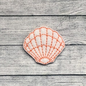 May include: A white seashell-shaped felt applique with orange stitching. The applique has a fan-like pattern and is placed on a gray wooden surface. The design is reminiscent of coastal decor.