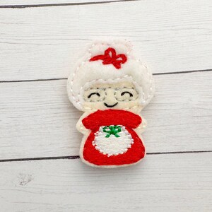 May include: A felt Mrs. Claus ornament with a white head and a red dress. The ornament has a smiling face with silver glasses, a red bow, and a green and white apron design. The edges are stitched with white thread.