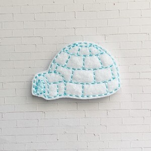 May include: A white felt igloo with light blue stitching. The igloo has a rounded dome and a small entrance. The stitching outlines the blocks of the igloo and adds a decorative element. The igloo is set against a white brick background.
