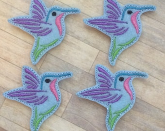 May include: Four felt hummingbird appliques. Each applique features a light blue body, purple wings, a pink throat, and a blue beak. Green and blue stitching outlines the hummingbird's shape. The appliques are arranged on a light brown wooden surface.
