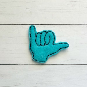 May include: A turquoise felt hand gesture, the shaka sign, with black stitched outlines. The felt is a solid color, and the stitching is neat and consistent. The background is a white, wooden surface.