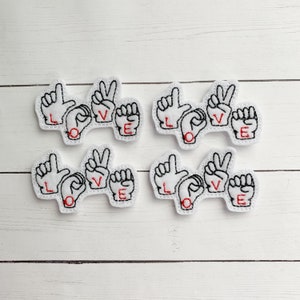 May include: Four white felt patches with the word "LOVE" spelled out in American Sign Language. Each patch features black embroidered outlines and red lettering. The patches are arranged on a white wooden surface.