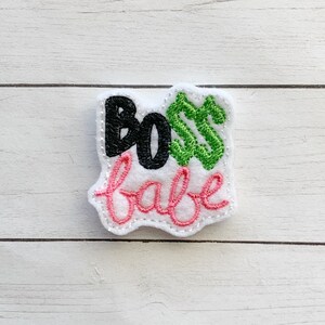 May include: A white felt patch with the words "BOSS babe" embroidered in black, green, and pink thread. The patch has a scalloped edge and is designed to be an accessory.