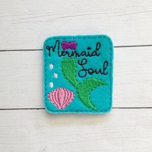 May include: A square teal felt patch featuring black embroidered text that reads Mermaid Soul. The design includes a green mermaid tail, a pink seashell, a small purple bow, and three white circular bubbles stitched onto the fabric surface.