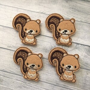 May include: Four embroidered squirrel patches. Each patch features a light brown squirrel with a dark brown tail and accents. The squirrel has a cream-colored embroidered belly and face, with black eyes and a black nose. The patches are on a light gray wooden surface.