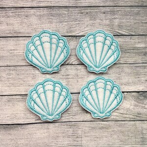 May include: Four embroidered seashell patches. Each patch features a white shell with a light blue outline and interior lines. The patches are arranged in a square on a wooden surface.