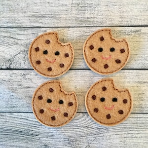 May include: Four felt cookie decorations with a bite taken out of each. The cookies are tan with brown chocolate chips and a stitched smiling face. The background is a light gray wooden surface.