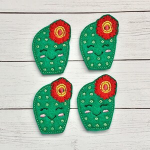 May include: Four green felt cactus appliques with smiling faces and red felt flowers on top. Each cactus has a yellow center in the flower.