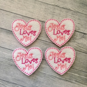 May include: Four white felt heart-shaped patches with pink stitching around the edges. Each patch has the words "Jesus Loves Me!" embroidered in pink and white.