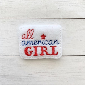 May include: A white rectangular patch with the embroidered text "all american GIRL" in red and blue thread. A red star is embroidered above the word "american". The patch is on a white wooden surface.