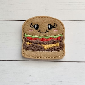 May include: A felt appliqué of a smiling hamburger with eyelashes, a green lettuce leaf, a red tomato, a brown patty, and a yellow cheese slice.