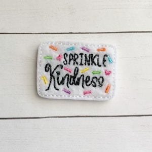 May include: A white felt patch with the words "SPRINKLE Kindness" embroidered in black script. Colorful embroidered sprinkles are scattered across the patch. The patch is rectangular with rounded corners.