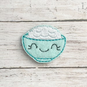 May include: A light blue felt patch in the shape of a bowl of rice with a smiling face. The rice is white, and the bowl has a teal outline and a teal embroidered border. The patch is on a light wood grain surface.
