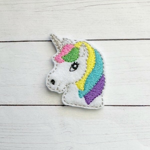 May include: A white felt unicorn patch with a silver horn and colorful mane. The mane features pink, green, yellow, turquoise, and purple stripes. The unicorn has a black eye and is outlined with gray stitching.
