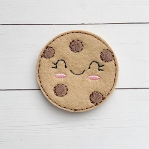 May include: A brown felt cookie with a smiling face and chocolate chips. The cookie is embroidered with a happy face and pink cheeks.