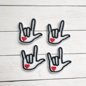May include: Four white patches embroidered with a black outline of a hand making the "I love you" sign in American Sign Language. Each patch has a small red heart in the center of the palm. The patches are arranged on a white wooden surface.